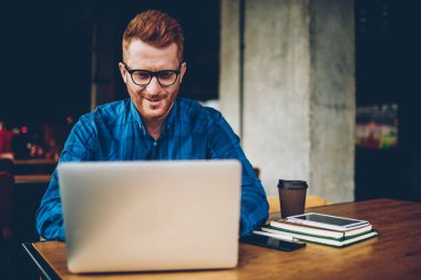 Neşeli erkek o kızıl saçlı yükleme ile profesyonel programı coorkin uzayda oturan modern dizüstü bilgisayarda. Zeki öğrenci üniversitede netbook üzerinde web sitelerinde iyi haber okuma gözlük