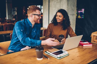 Gülümseyen erkek öğretmen dizüstü bilgisayar coworking içinde oturan neşeli Afro-Amerikan öğrenci çalışma için yeni konu açıklama Kızıl saçlı. İki hipsters yeni proje işbirliği sırasında tartışıyor