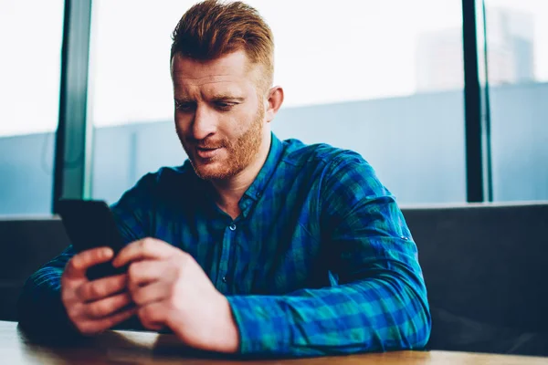 Male blogger with red hair charcking email and chatting online on smartphone connected to 4G internet sitting in coworking space.Young man in casual shirt reading incoming notification on cellular