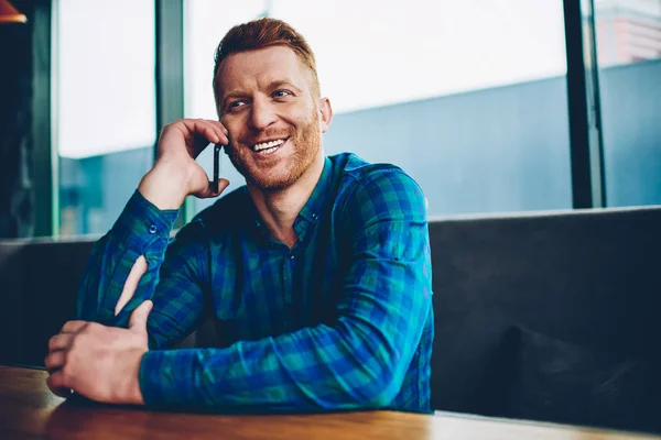 Happy young man with red hair satisfied with good mobile connection in roaming while talking with friends on smartphone device.Positive male communicating on cellular resting in coworking space