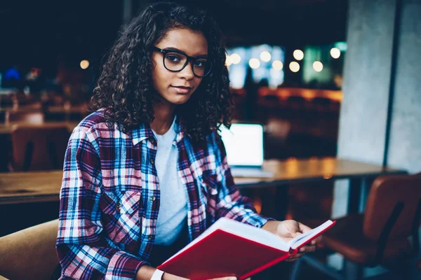 Kıvırcık saçlı ve kitap okuyan ile Kitaplığı'nda otururken kameraya bakarak gözlük güzel akıllı Afro-Amerikan öğrenci portresi. Zeki koyu tenli teen elinde ders kitabı ile