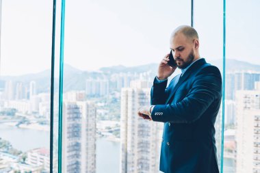 Düşünceli erkek avukat smartphone hakkında konuşurken kol saati görünümlü iş toplantı düzenlemek. Kendine güvenen gurur CEO'su zaman planlama Konferansı ayakta Office yakınındaki pencere denetimi