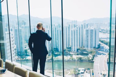 Ofiste ayakta ve metropolis harika manzarasını penceresinden bakarak ssuccessful iş projesi sonuçlarını smartphone aygıt yolu ile söz formal giyim erkek girişimci arkadan görünüm