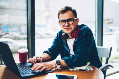 Half length portrait of of positive skilled freelancer in spectacles creating presentation for customer using laptop.Handsome happy hipster guy looking at camera while downloading files from network clipart