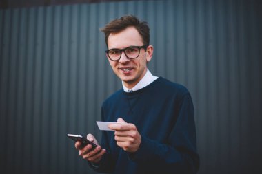 Yarım uzunlukta trendy Eyewear açık havada eğlence keyfini gülümseyen yakışıklı erkek öğrenci portresi. Kamera tutarken bakarak neşeli hipster adam ziyaret kart ve akıllı telefon yolu ile online rezervasyon