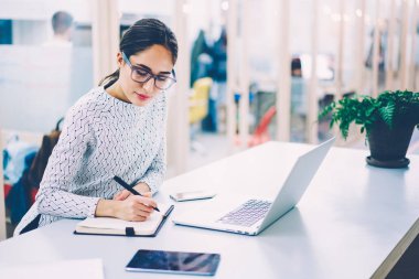 Dalgın kadın dizüstü dizüstü bilgisayar ile çalışma evinde otururken yazma çalışma programı planlama Eyewear kadın İdari İşler Müdürü notlar bilgi netbook üzerinde göz.