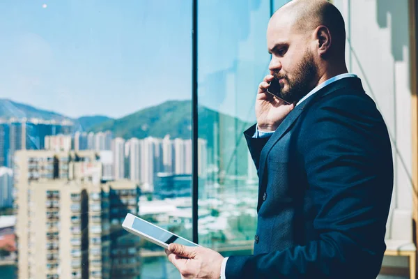 Side view of confident proud ceo dressed in formal apparel talking about banking transaction via smartphone while checking account on modern tablet standing near glass window with high skyscrapers on background