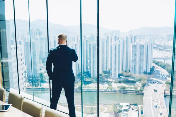 Arkadan görünüşü işadamı ciddi telefon sohbet ederken elini cebine tutarak ve güzel metropolis sahne ile pencereden dışarı bakıyor. Gurur CEO'su smartphone ayakta Office konuşurken