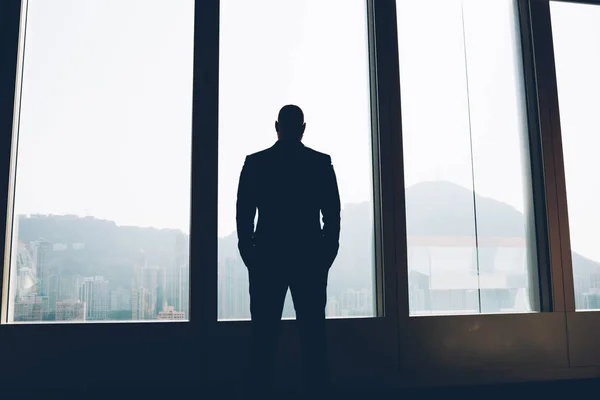 Back view of male proud ceo dressed in elegant wear looking out of window while thinking on working issues after hard day.Silhouette of businessman standing indoors in office of own company