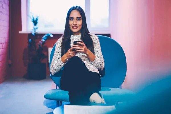 Positive brunette woman using mobile phone for networking on work break in office,young hipster girl chatting with friend online on smartphone share multimedia files sitting at share indoors