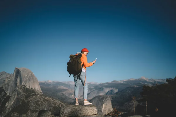 年轻女性旅行者与背包站立在岩石悬崖持有细胞得到新的消息使用良好的移动连接 女人流浪通过应用智能手机和网络获得山顶上 — 图库照片