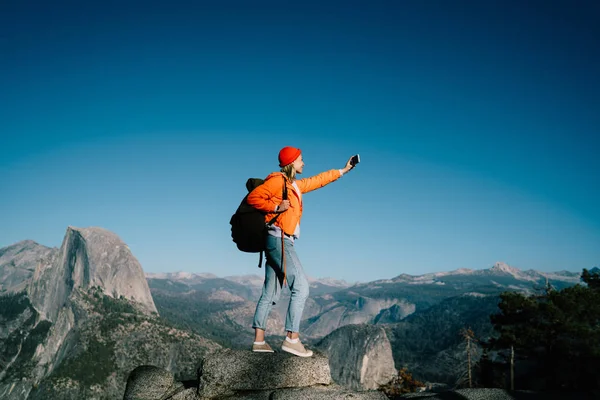Young female travel blogger taking selfie on smartphone camera during hiking tour in mountain,girl wanderlust with backpack posing for photo on telephone standing on breathtaking scenery on rock peak