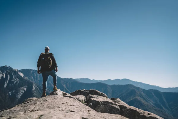 Back view of male explorer with backpack standing on rock cliff looking at breathtaking scenery of nature landscape, hipster guy wanderlust getting to mountain peak on hiking tour in summer