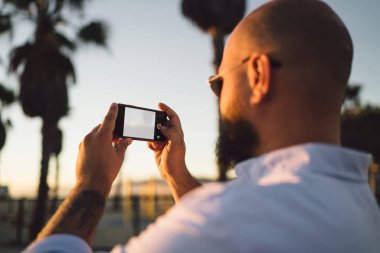 Erkek blogger fotoğraf çekmek için sahte ekranı ile smartphone kullanarak görünümünü sırt ve ağlarda, modern cep telefonu fotoğrafları yaz tatillerini akşamları sırasında yapma hipster adam pay