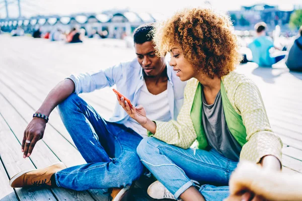 Afro american couple in love using modern smartphone for watching movie online sitting outdoors in city park,pretty dark-skinned girl with curly hair viewing photos on mobile with boyfriend for share