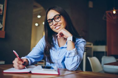 Optik gözlük Not Defteri'nde şık giyim taslağını çizerken kameraya gülümseyen güzel neşeli tasarımcısında yarım uzunluk portre. Not Defteri'nde yapma olumlu çekici öğrenci