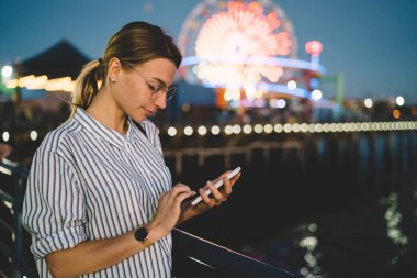 Genç kadın blogger paylaşmak içerik ağlarında smartphone Santa monica İskelesi akşam, hippi kız manifatura mesaj beach eğlence parkı parlayan ışıklar üzerinde duran cep telefonu üzerinde yeniden oluşturmak yoluyla