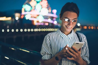 Hippi kız GÖRÜNTÜLENMESI akşam eğlence eğlence parkı, neşeli kadın turist parlak dönme dolap gece adam üzerinde cep telefonu duran yeni iletide okuma zevk blog gülümseyen