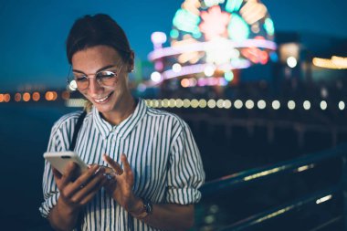 Hippi kız GÖRÜNTÜLENMESI akşam eğlence eğlence parkı, neşeli kadın turist parlak dönme dolap gece adam üzerinde cep telefonu duran yeni iletide okuma zevk blog gülümseyen
