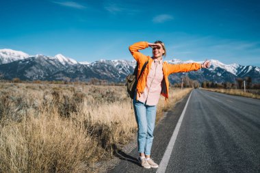 Asfalt karayolu, neşeli kız yolculuk tutkusu autostop hafta sonları Americ vahşi doğada keşfetmek harcama tarafından seyahat üzerinde gösteren iç bekleyen araç gülümseyen genç kadın otostopçu tam uzunlukta