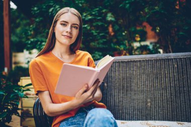 Kitap kopyalama alanı alanı reklam alanı için yakındaki tahta bankta ile istirahat ederken kameraya bakarak akıllı genç kadın yarım uzunluk portre. Yetenekli erkek edebiyat eğitimi