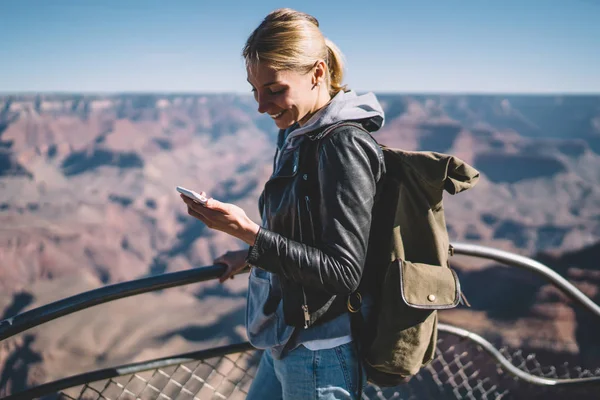 Smiling female explorer reading message on telephone satisfied with mobile connection during hiking tour,cheerful  girl wanderlust using cellular chatting standing on viewpoint in Grand Canyo