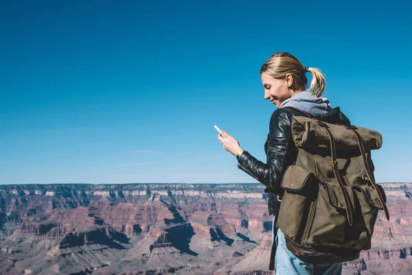 Smiling female explorer standing on breathtaking Grand canyon scenery reading message on smartphone happy about good connection, girl wanderlust with backpack checking cellphone mail in roaming