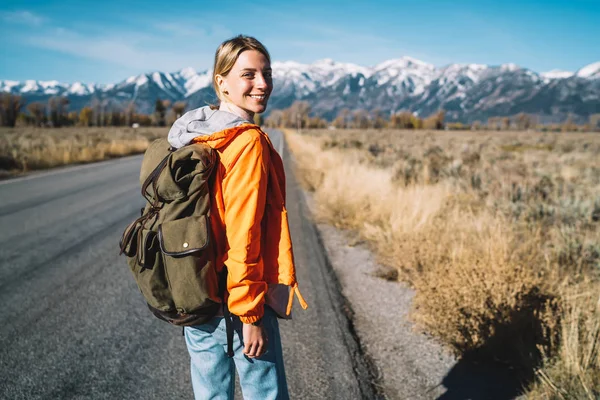 Sırt çantası ile neşeli kadın otostopçu yarım uzunluk portre ile hafta sonu gezisi, ufuk zevk yolculuk adventur dağda tanışmadan asfalt yolda yürüyen kız yolculuk tutkusu gülümseyen heyecan