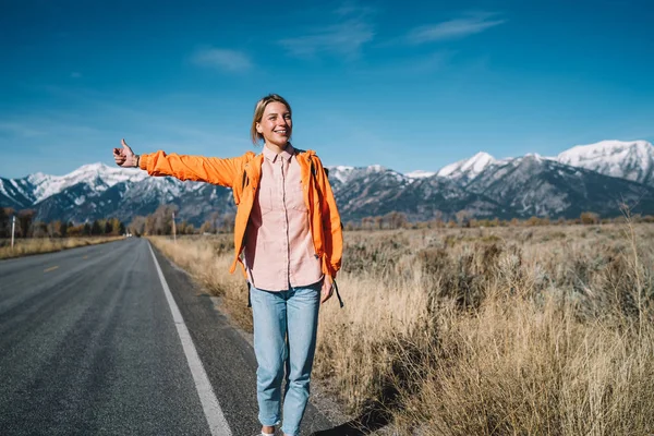 Cheerful female explorer excited with adventures during trip standing on asphalt road hitchhiking, happy girl wanderlust waiting for car to stop on highway getting to destination in national par