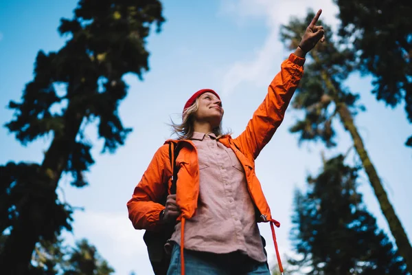 Positive young female explorer enjoying hiking trails in wild wood environment showing direction, smiling hipster girl wanderlust in orange jacket pointing standing in forest discovering natur