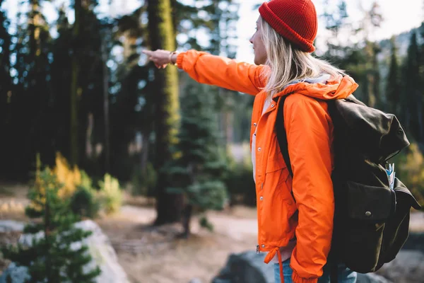 Back view of female explorer pointing on trail in forest discover wild environment on active weekend tour, girl wanderlust with touristic rucksack showing direction raising hand hiking in wood