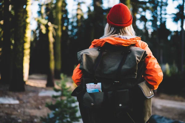 Back view of female traveler in colourful orange jacket recreating during hiking tour in forest, girl wanderlust with touristic rucksack and backpack explore wild environment of wood on active res