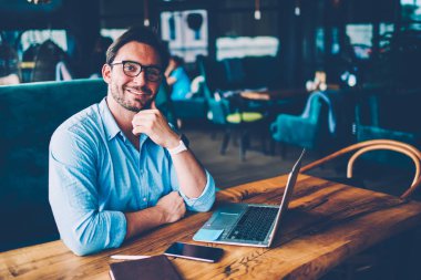 Şık gömlek ve optik gözlükler uzaktan netbook oturma tanıtım alanı kafede, çalışırken kameraya gülümseyen başarılı sakallı yakışıklı işadamı portresi yarım uzunluk