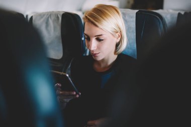 Güzel bonde kadın yolcu smartphone online uçak birinci sınıfta rahat koltuğunda oturan iş gezisi sırasında kablosuz internet bağlantısı ile iletişim e-posta kontrol
