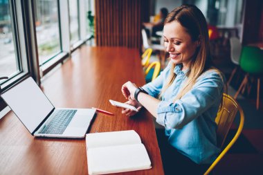 Sarışın saçlı boş ekran alanı'nda şık coworking ile textboo ve dizüstü bilgisayar başında oturan akıllı sms mesaj yollama zamanı kontrol smartwatch bakarak mutlu öğrenci