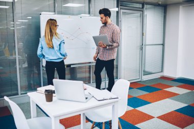 Kadın profesyonel öğretmen flipchart üzerinde modern netbook ile yetenekli öğrenci el çizilmiş diyagramı açıklayan. Nitelikli koç özel ders kurulu üniversitede yakın duran hipster adam için iletken