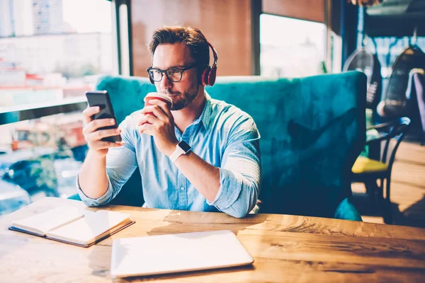 沉思胡子企业家在现代耳机听音乐和喝可口的咖啡 同时阅读财经新闻互联网网站通过4G 连接到智能手机坐在咖啡店 — 图库照片