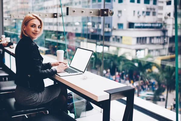 微笑的时尚年轻女子的肖像坐在现代笔记本电脑与空白屏幕区域和做远程工作 在咖啡馆为自由职业者的4G 互联网连接 — 图库照片