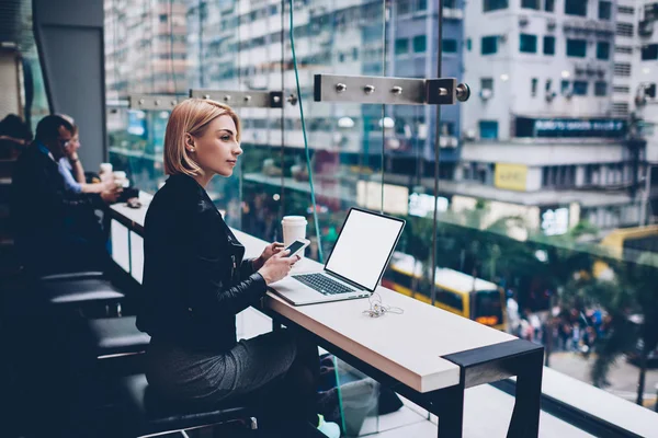 Thoughtful female blonde blogger with short haircut looking out of window with cityscape and updating profile on smartphone sitting at netbook with blank copy space screen for advertising information