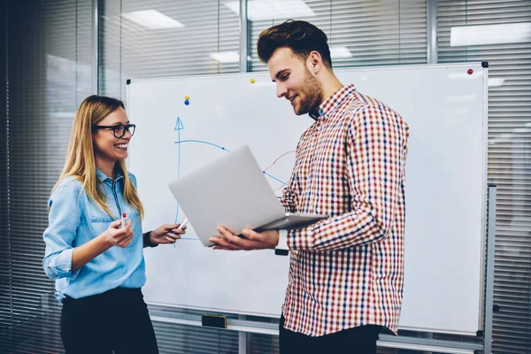 Positive hipster student showing funny presentation to smiling female teacher on laptop computer standing near flipchart.Successful male tutor explaining theme of video lesson to young woman