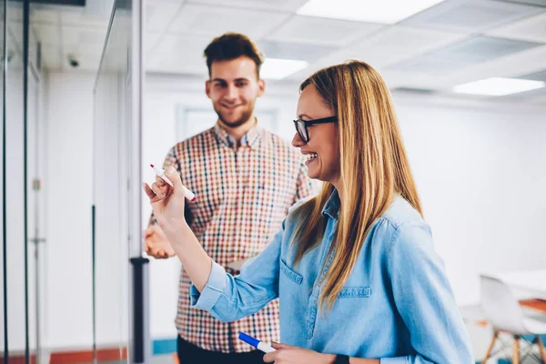 Positive young male coach dictating information to skilled student for writing .Cheerful student with blonde hair laughing while drawing digram during interesting lesson with successful coach