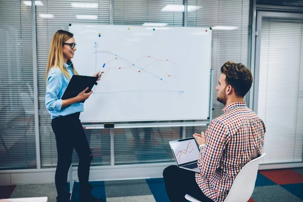 Professional female coach with marker in hand pointing on flipchart with diagram and explaining theme to skilled student sitting with laptop computer during private lesson in university