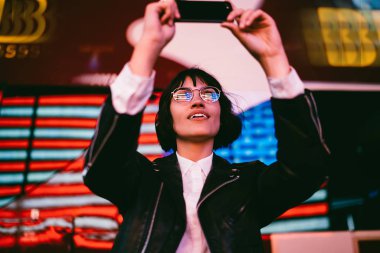 Esmer hippi kız New York'ta gece yürüyüş zevk ve Smartphone için blog yayında fotoğrafları yapma Eyewear hayran. Heyecanlı kadın şık aşınmaya neon aydınlatma ile video çekim