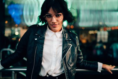 Yarım uzunluk portre çekici esmer hippi kız gözlük içinde boş zaman akşam içinde kentsel ortamda arka plan bulanık fotoğraf makinesi ayakta bakarak rahat şık kıyafet giymiş