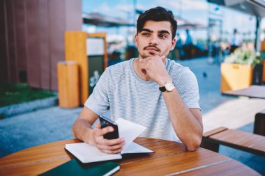 Kendi web sitesi ve yayın için yeni fikirler geliştirme düşünceli erkek blogger düşünme. El ile eğitim ders kitapları açık havada terracce okul sırası vasıl oturan smartphone ile durulması öğrenci