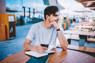 Düşünceli sakallı blogger düşünme üzerinde smartphone ile el ahşap masaüstü ders kitapları ile oturmuş. Web sitesinde yayın hikayesi için fikirler. Düşünceli öğrenci ile telefon planları üzerinde durulması