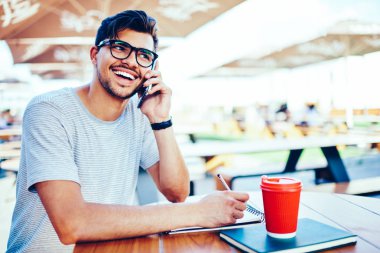 Söz ve ödev yapmak gitmek için ders kitabı ve kahve ile tahta masada oturan smartphone arkadaş telefon görüşme sırasında komik hikaye ile gülüyor gözlük olumlu hipster öğrenci