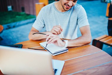 Croped görüntü otururken ahşap masa ile modern dizüstü bilgisayar, kablosuz internet bağlantısı ve Not defteri ile notlar için zaman yönetimi için smartwatch bakarak başarılı erkek öğrenci