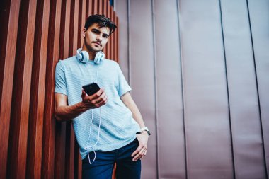 Yakışıklı hipster adam akıllı telefon elinde tutarken kameraya bakarak rahat şık kıyafeti portresi. Trendy erkek meloman telefon ayakta publisity alanı yakınında bağlı kulaklık ile giyinmiş