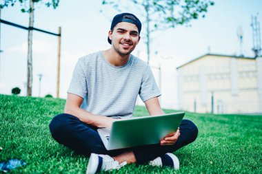 Neşeli hipster adam şık Cap kamera yükleme dosyasını Web sitesinden Park yeşil çimenlerin üzerinde oturan netbook üzerinde iken gülümseyen portresi. Kampüs içinde açık havada dinlenme dizüstü bilgisayar ile pozitif öğrenci